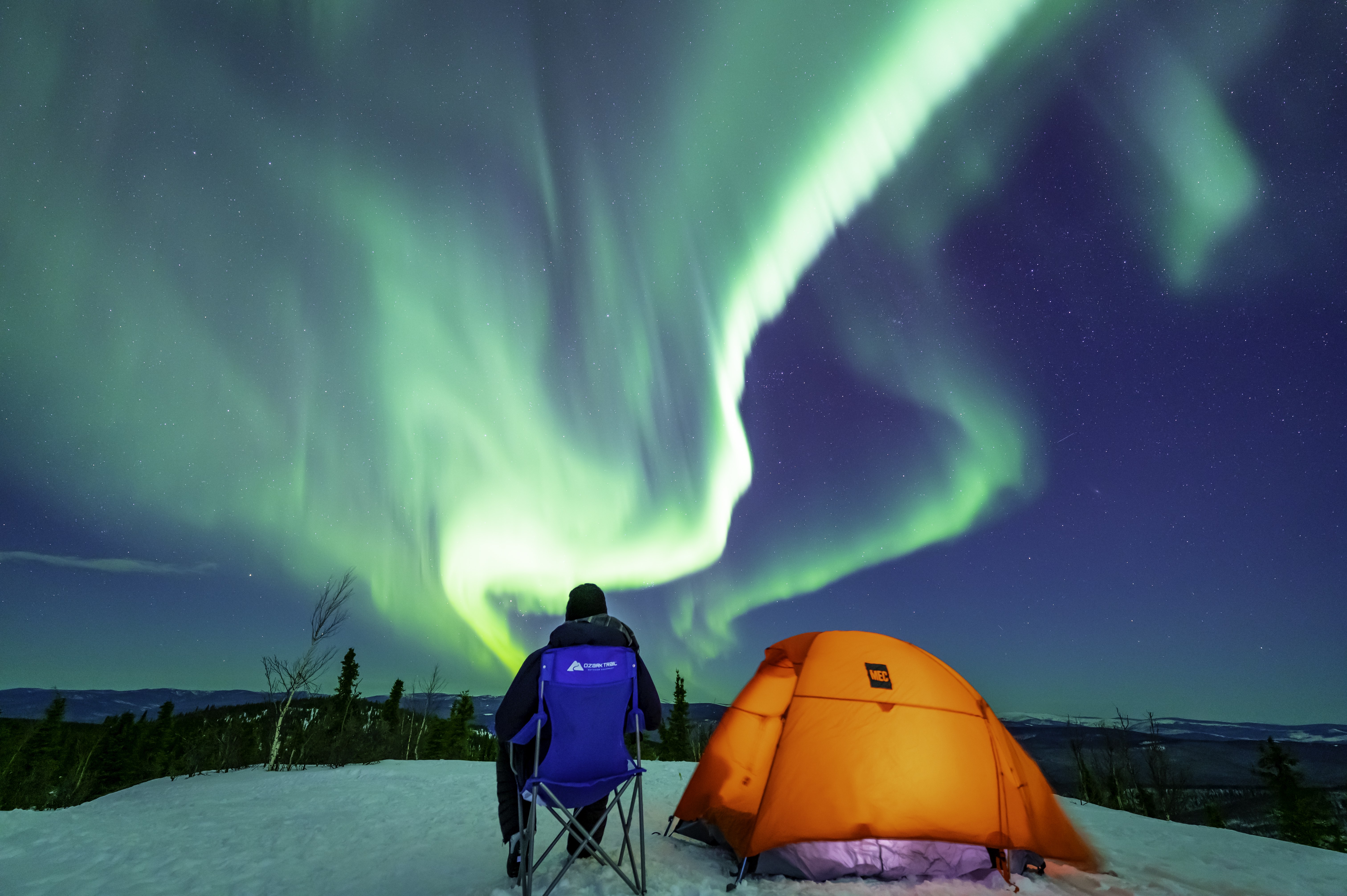 Aurora above a snowy Alaskan landscape, with a camper and an orange tent beneath the night sky.